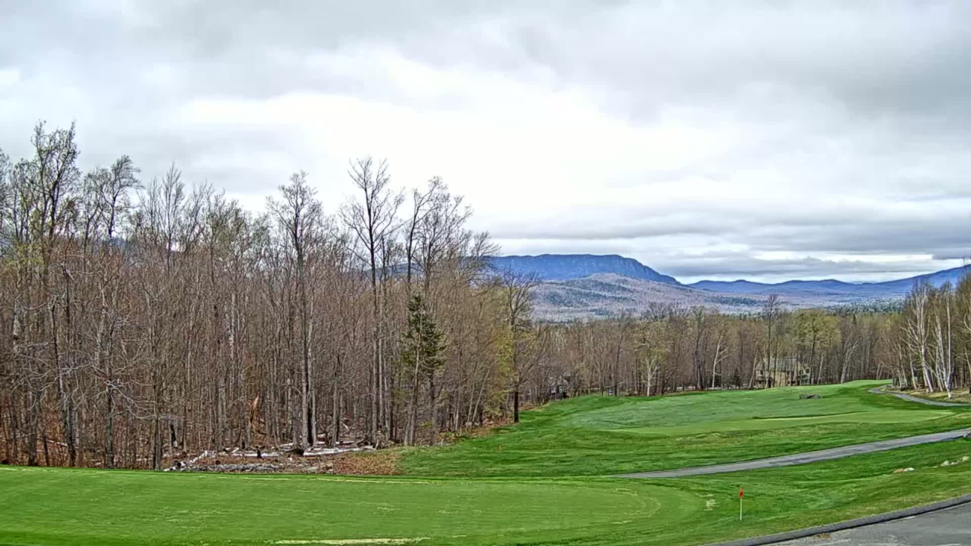 A golf course fairway stretches toward a line of trees and distant mountains under a cloudy sky.