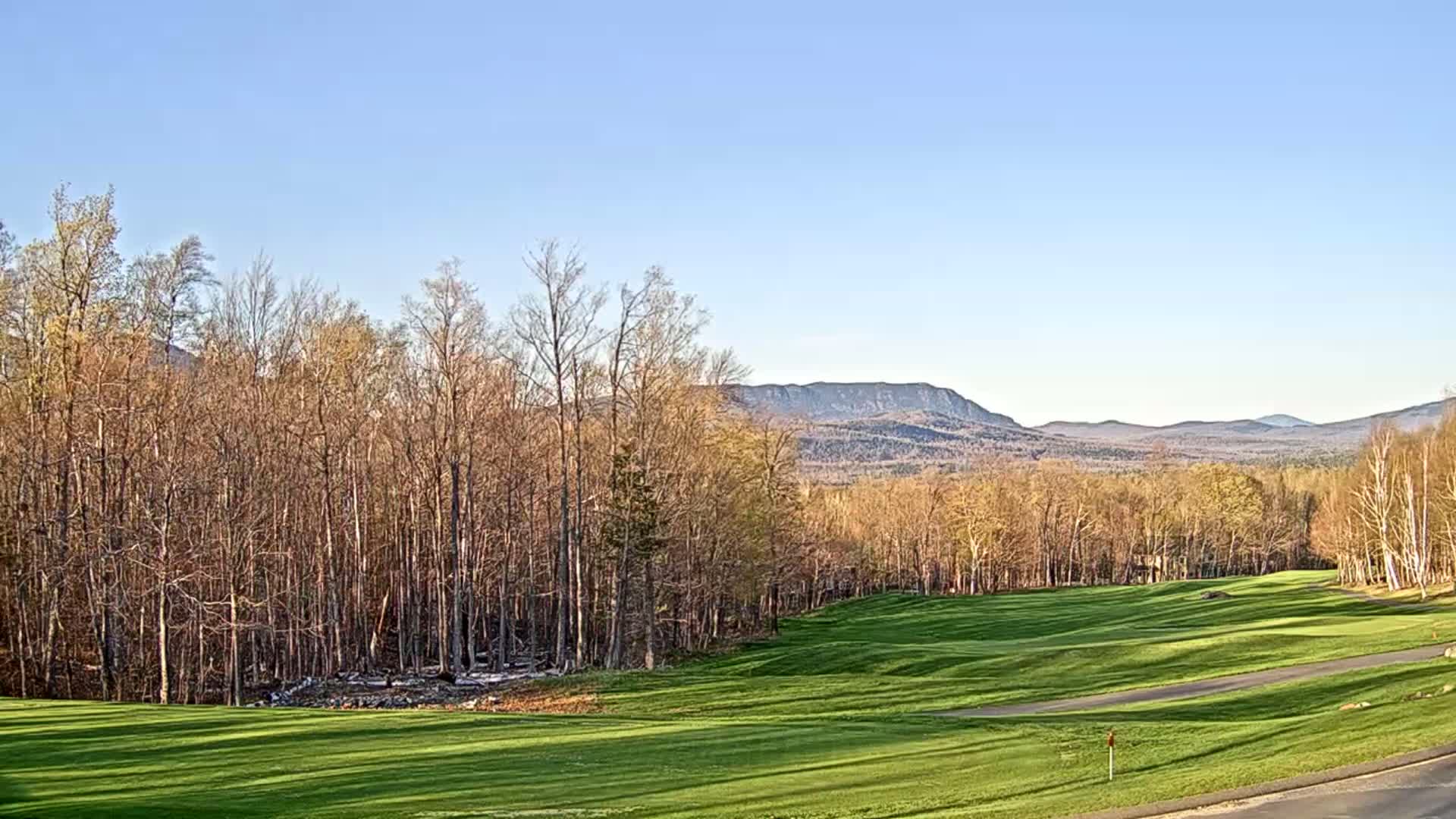 A sunny day reveals a golf course nestled among bare trees, with distant mountains in the background.
