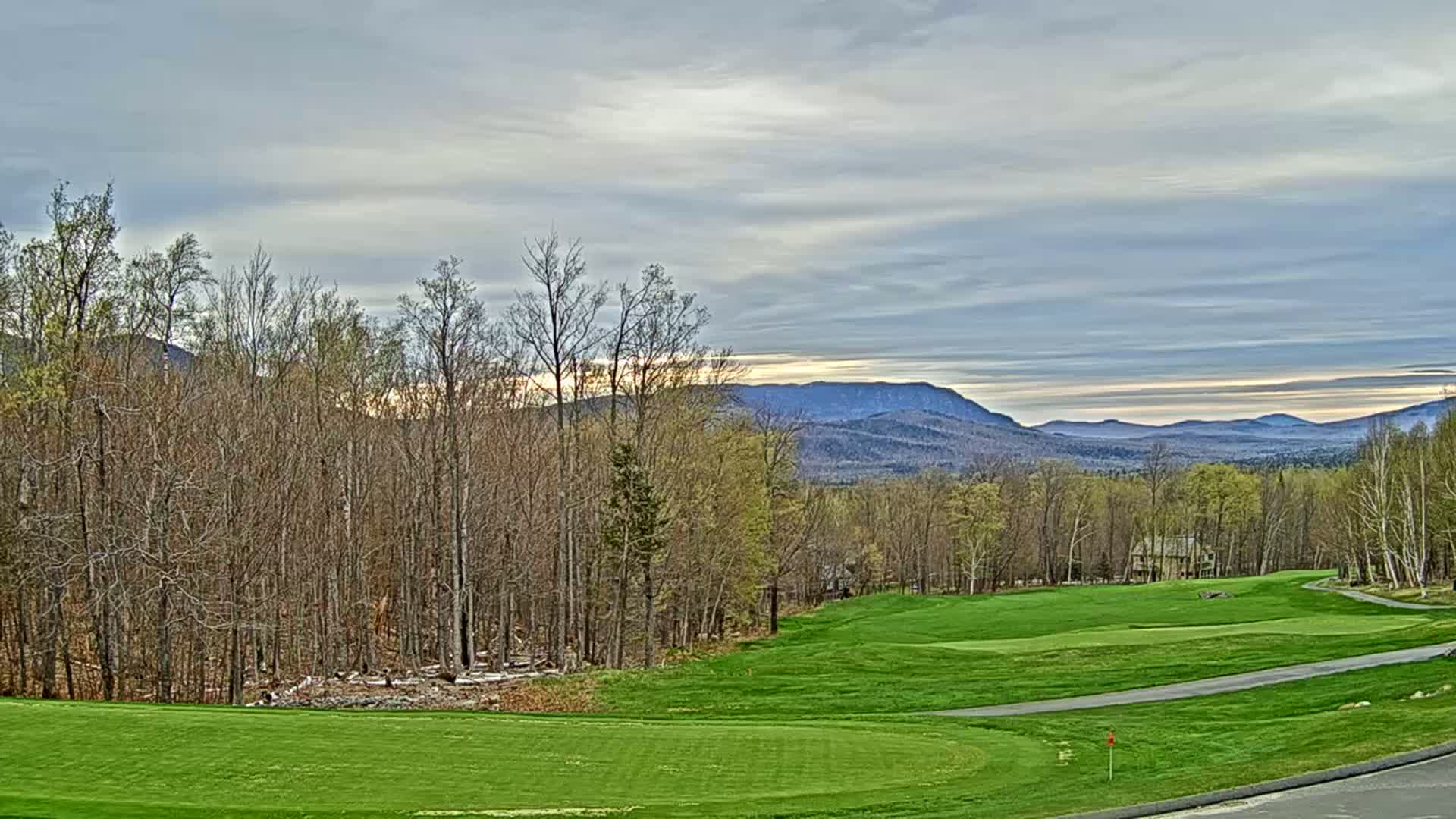 A golf course fairway stretches toward a line of trees and distant mountains under a partly cloudy sky.
