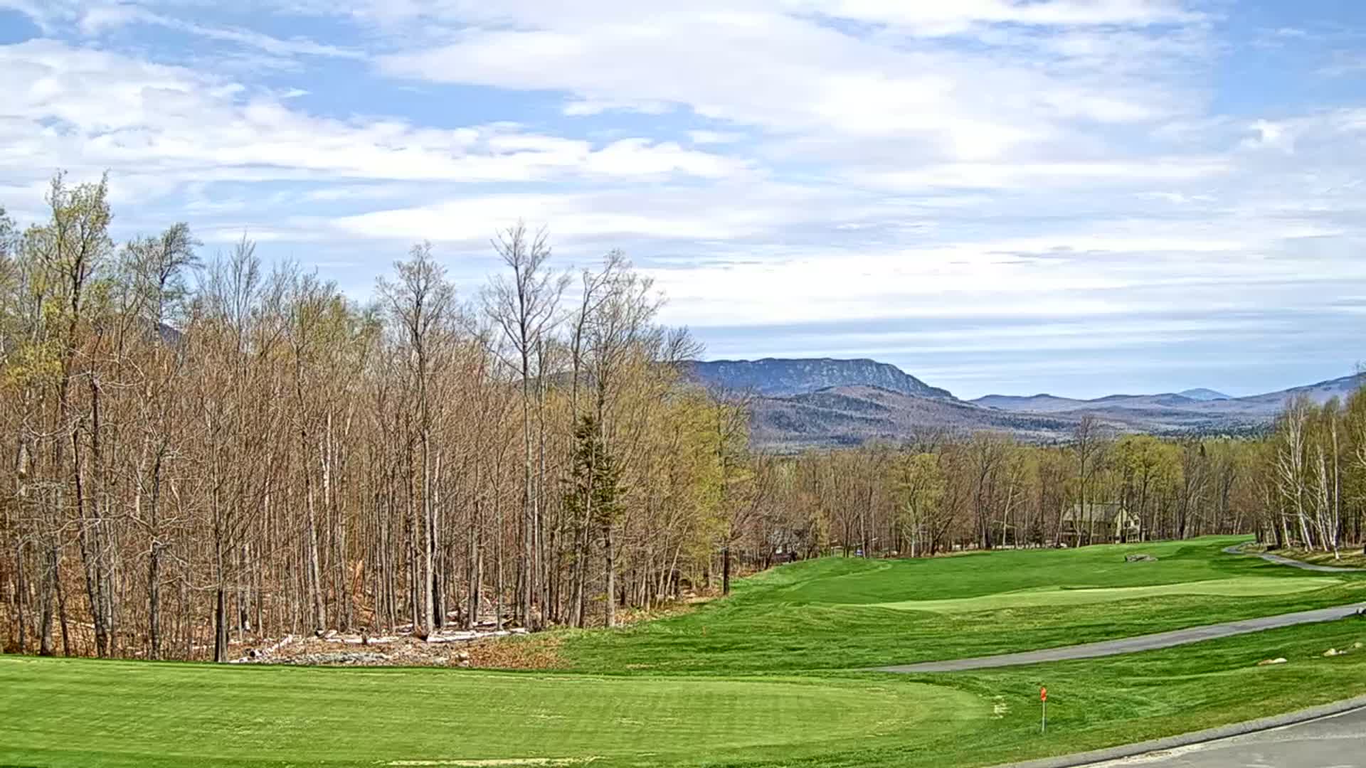 A partly cloudy sky overlooks a golf course bordered by a deciduous forest and distant mountains.