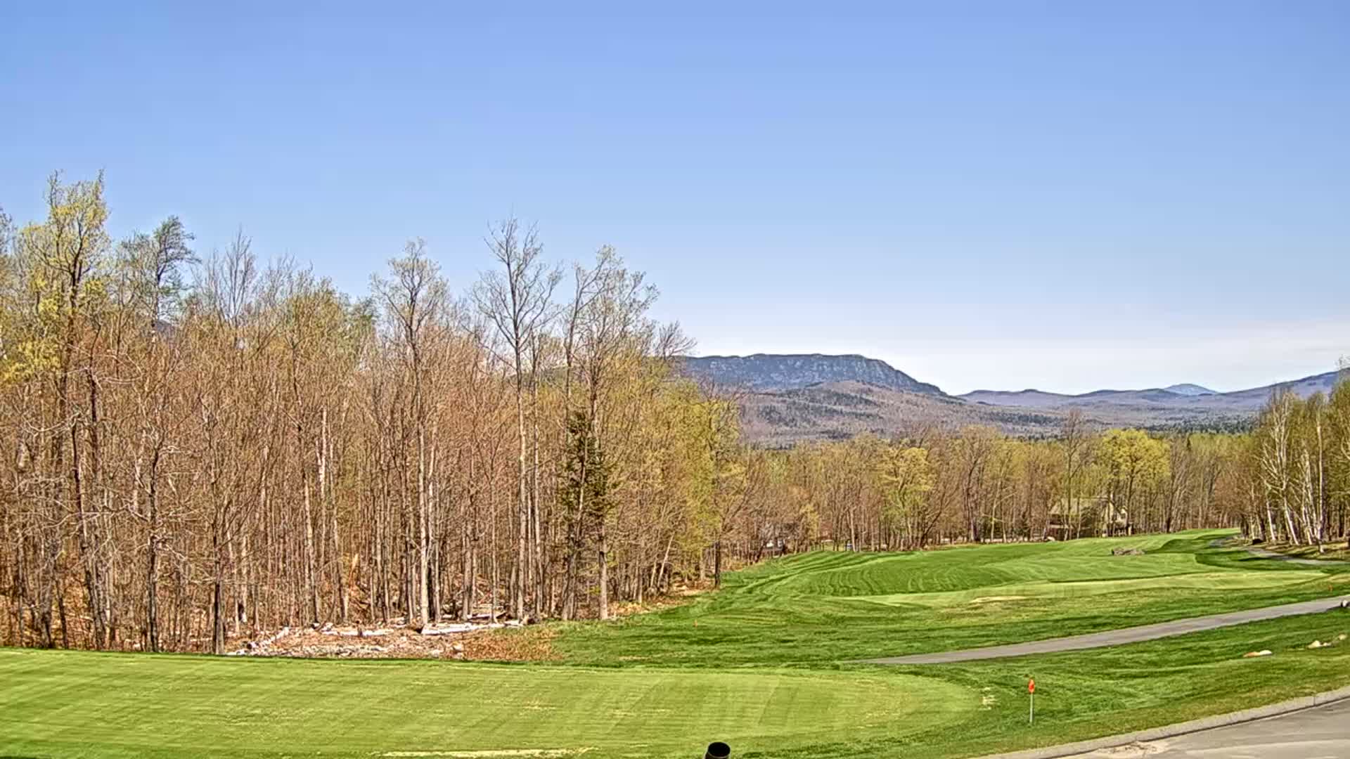 A golf course with a green fairway and trees stretches towards distant blue mountains under a clear blue sky.