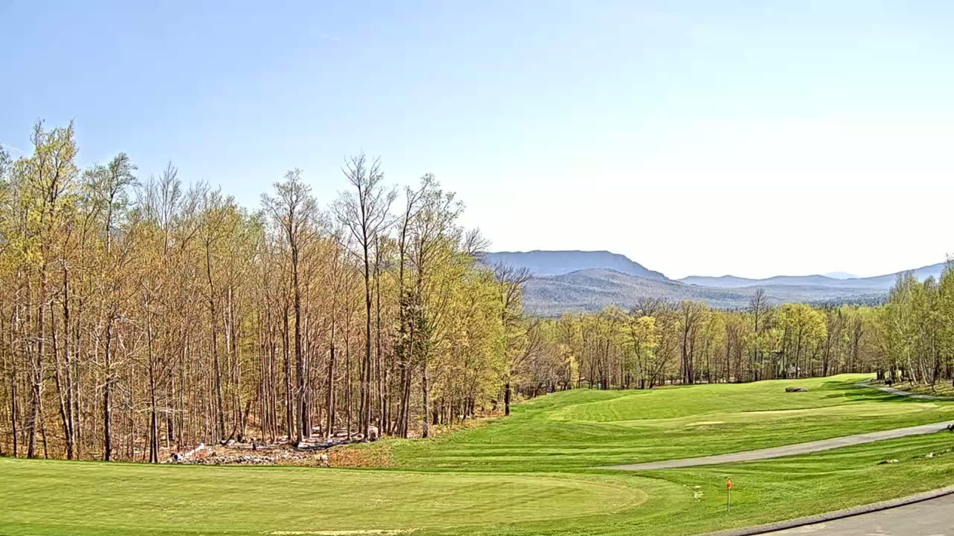A golf course fairway stretches towards a background of trees and distant, hazy mountains under a clear, bright sky.