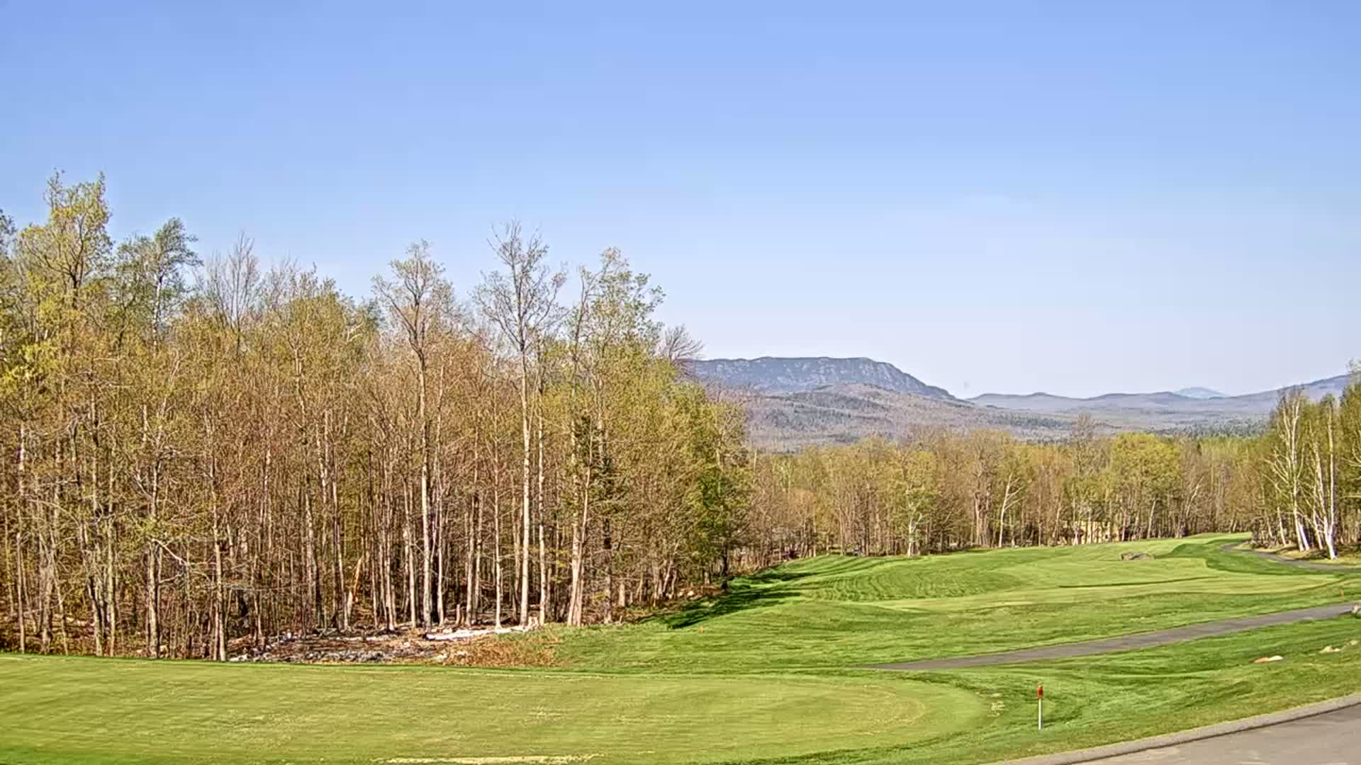 A scenic golf course with a green fairway, bordered by deciduous trees, stretches towards distant, hazy mountains under a clear blue sky.