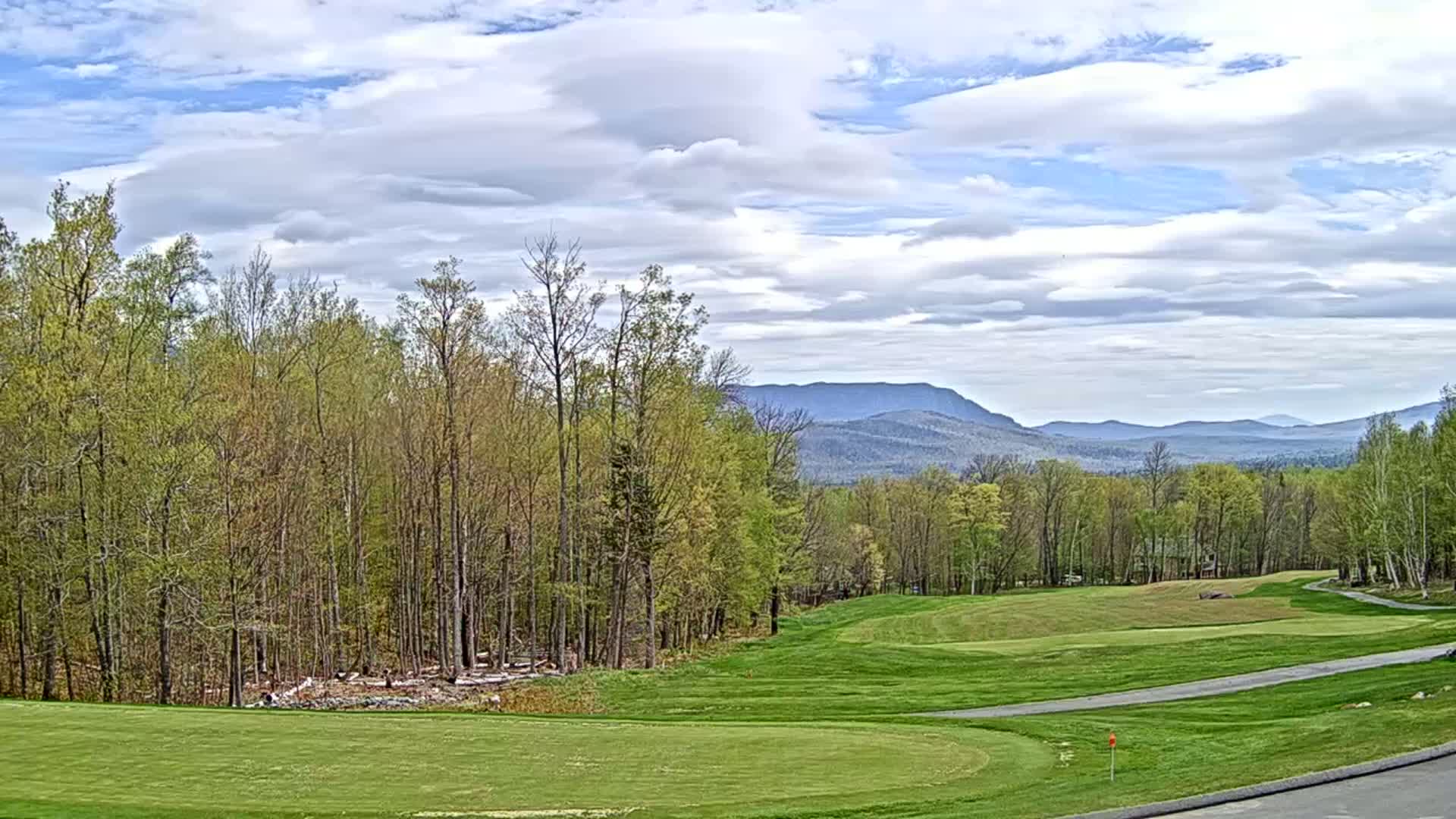 A partly cloudy sky overlooks a golf course nestled in a forest with distant mountains in the background.