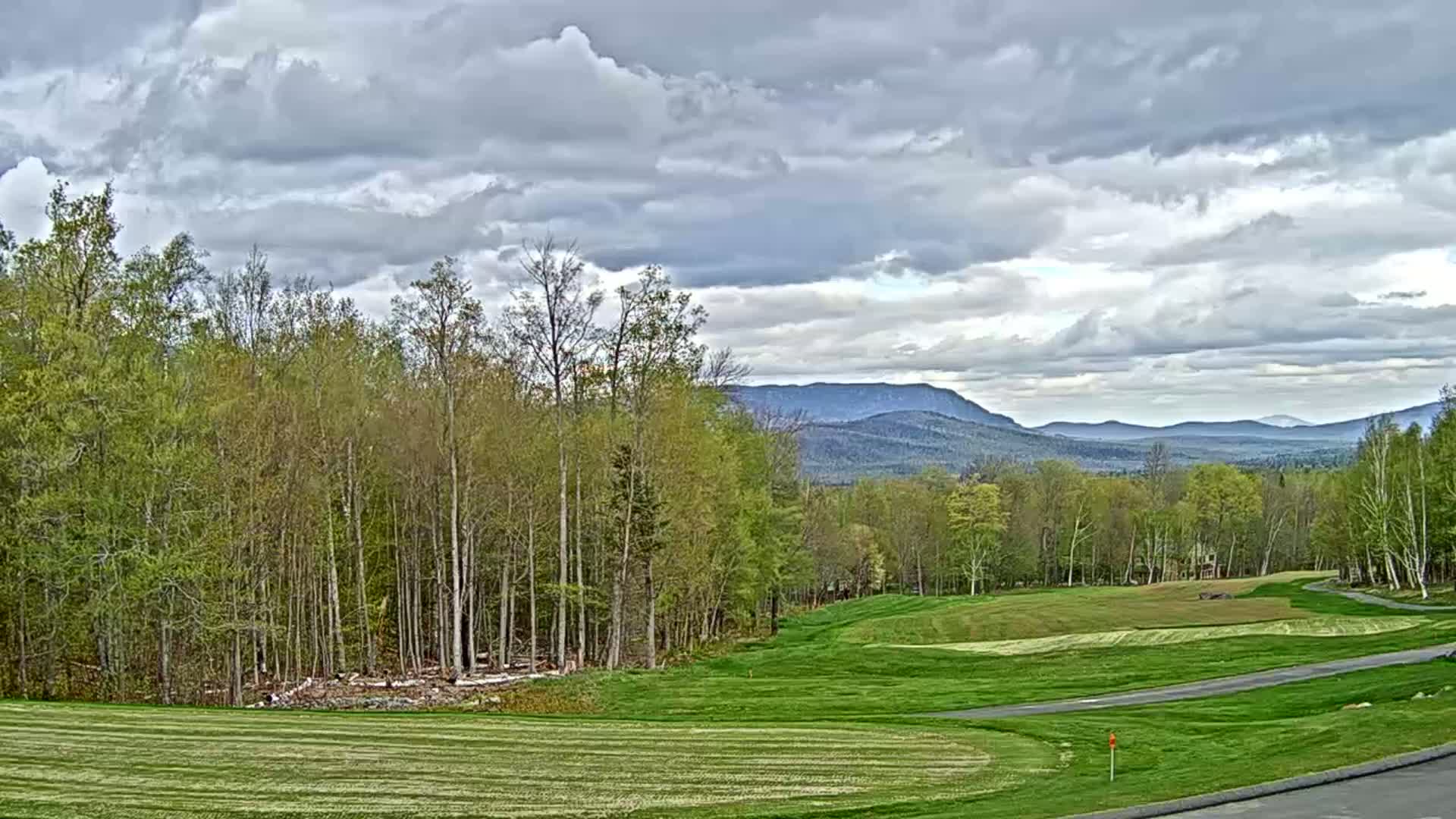 A partly cloudy day reveals a golf course bordered by a forest, with distant mountains visible under a mostly cloudy sky.