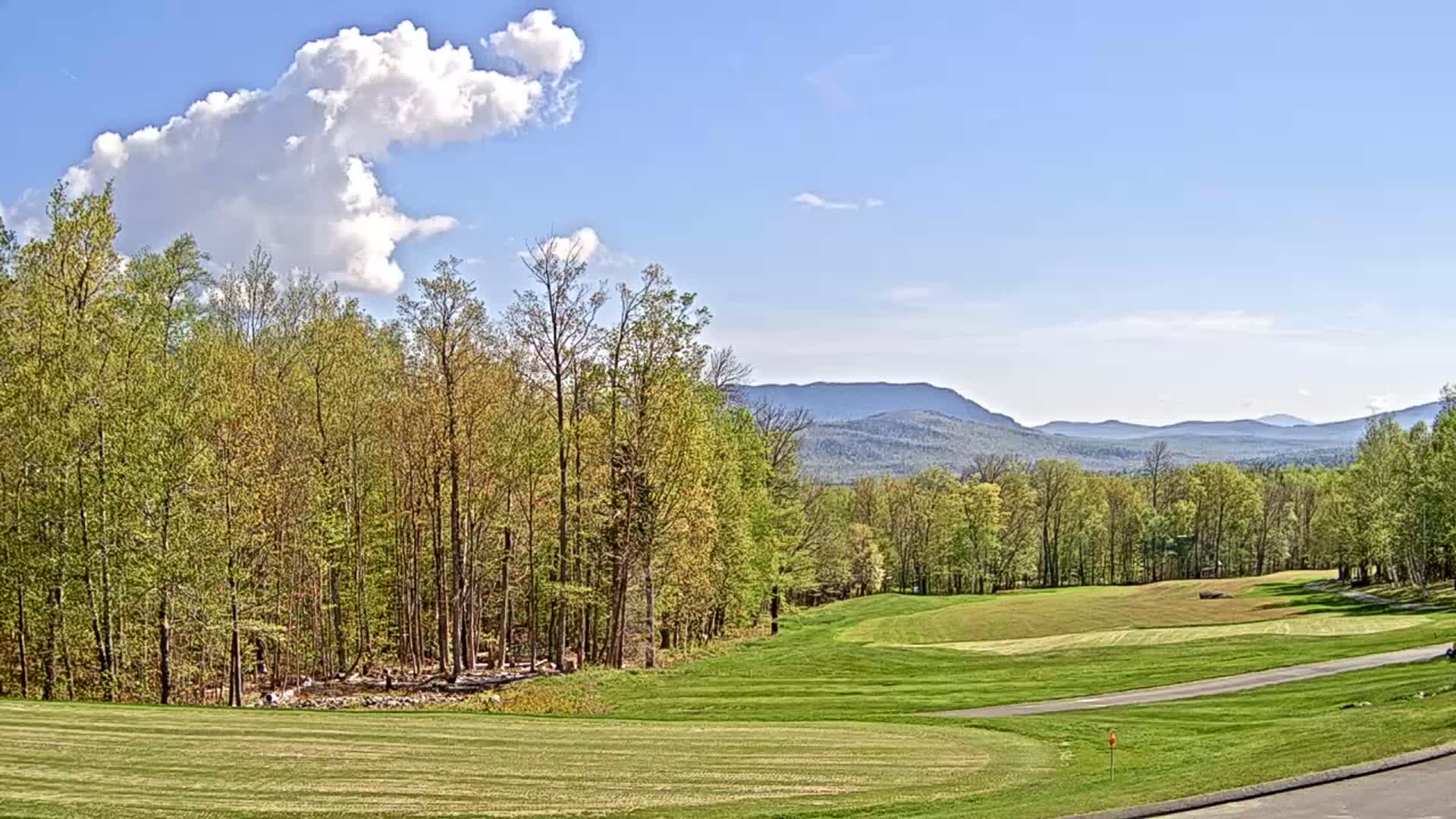 A partly cloudy blue sky overlooks a golf course nestled amongst a line of deciduous trees, with distant mountains visible on the horizon.