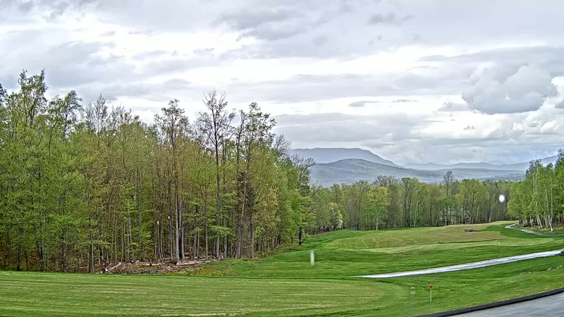 A golf course with a lush green fairway stretches out before a line of deciduous trees, under a mostly cloudy sky with distant mountains visible in the background.