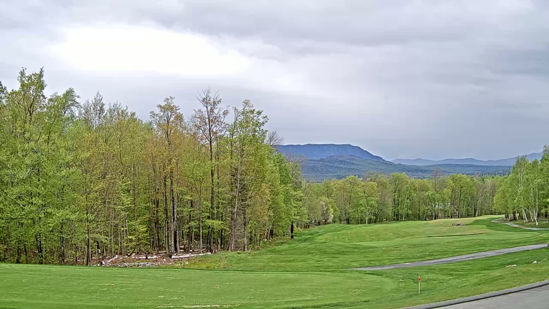 A golf course with green fairways and trees in the foreground, backed by a range of mountains under a cloudy sky.
