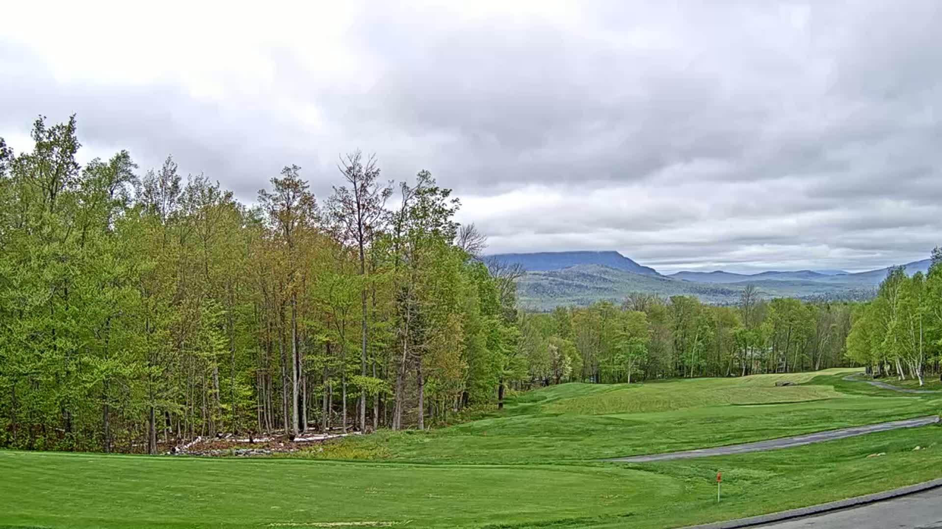 A golf course with green fairways and trees, overlooks a distant mountain range under a cloudy sky.
