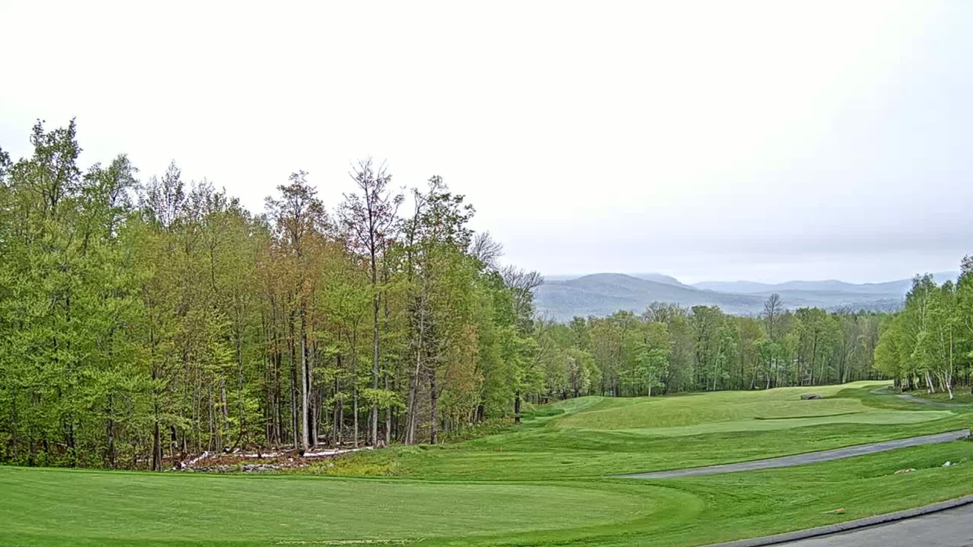 A golf course with green fairways and a wooded backdrop overlooks distant, hazy mountains under an overcast sky.