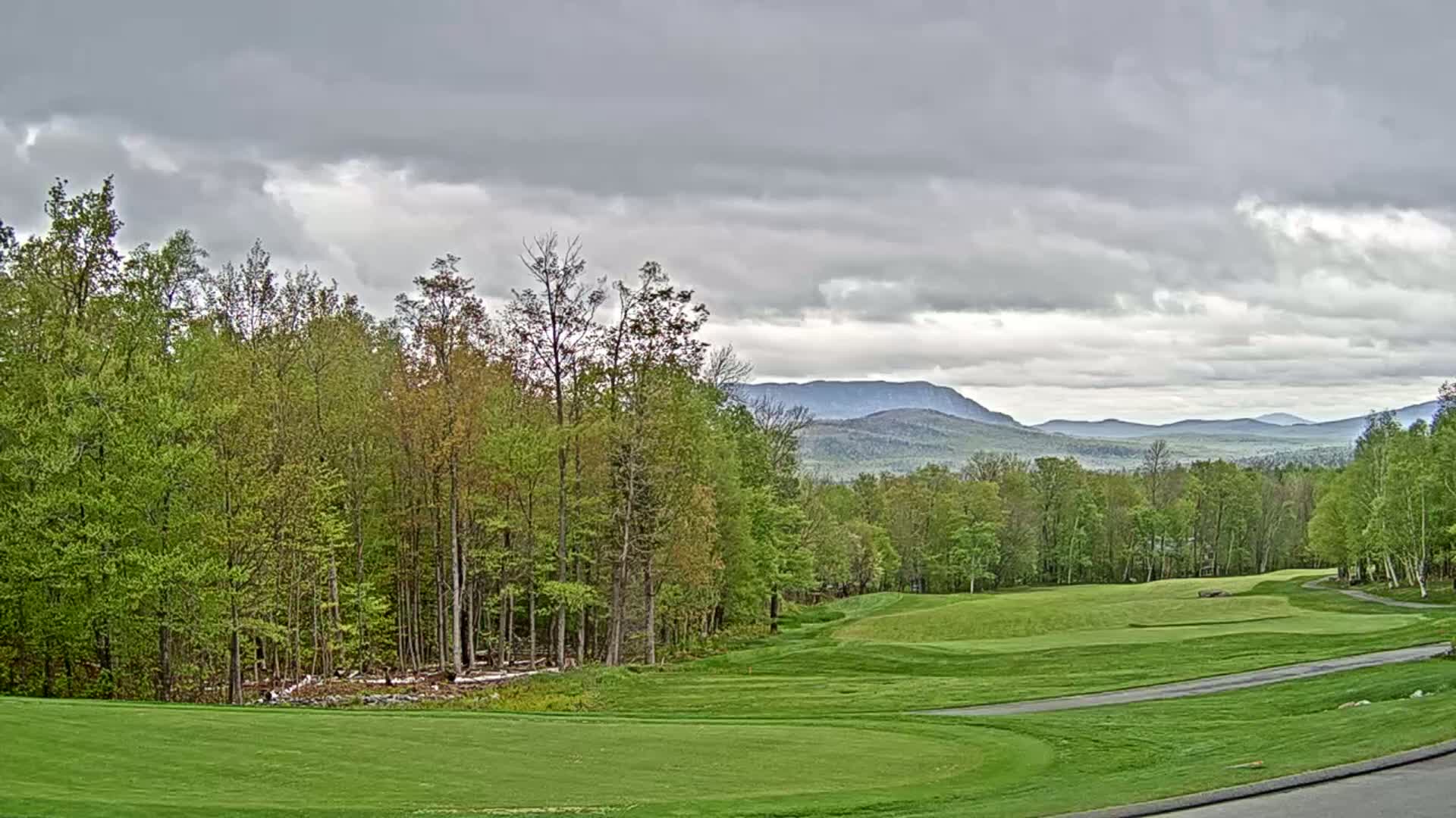 A golf course fairway and green stretch out toward a range of distant mountains under a cloudy sky.