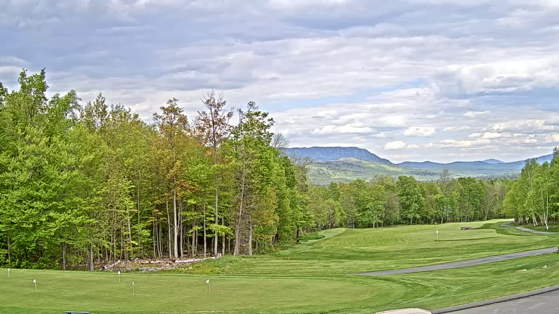 A golf course fairway and green are situated amongst lush green deciduous trees, with distant blue mountains visible under a partly cloudy sky.