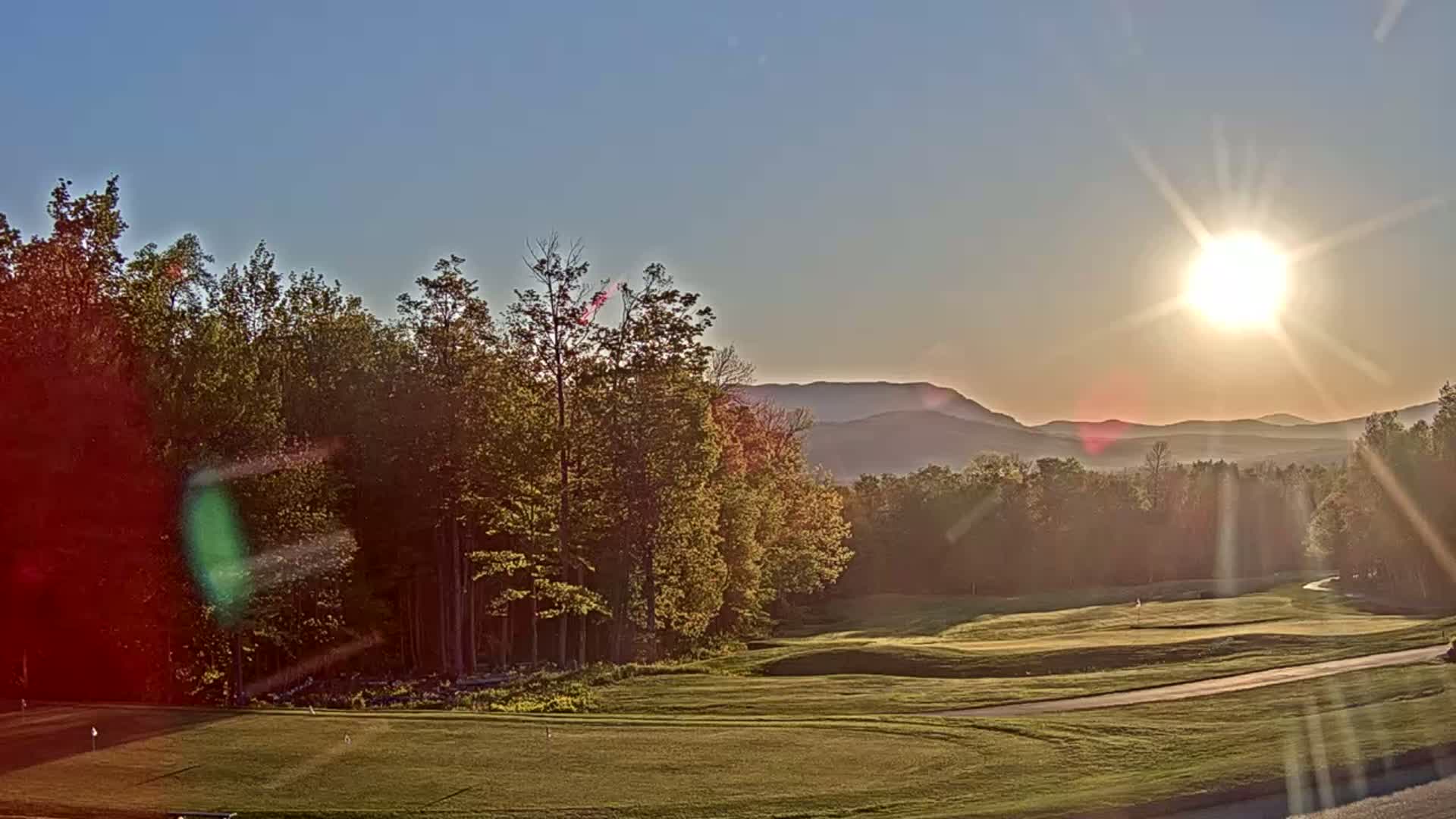 A golf course nestled among trees, with a bright sun setting behind distant mountains under a clear sky.