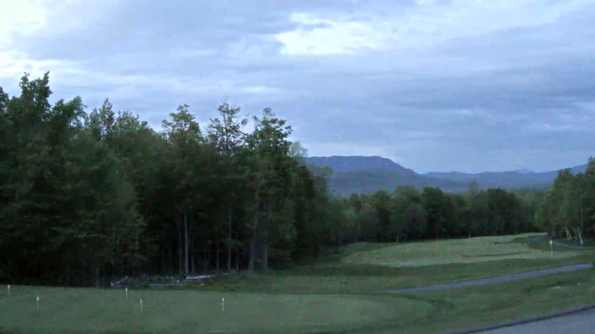 A golf course with green fairways and a line of trees in the foreground, overlooking a range of distant mountains under a cloudy sky.