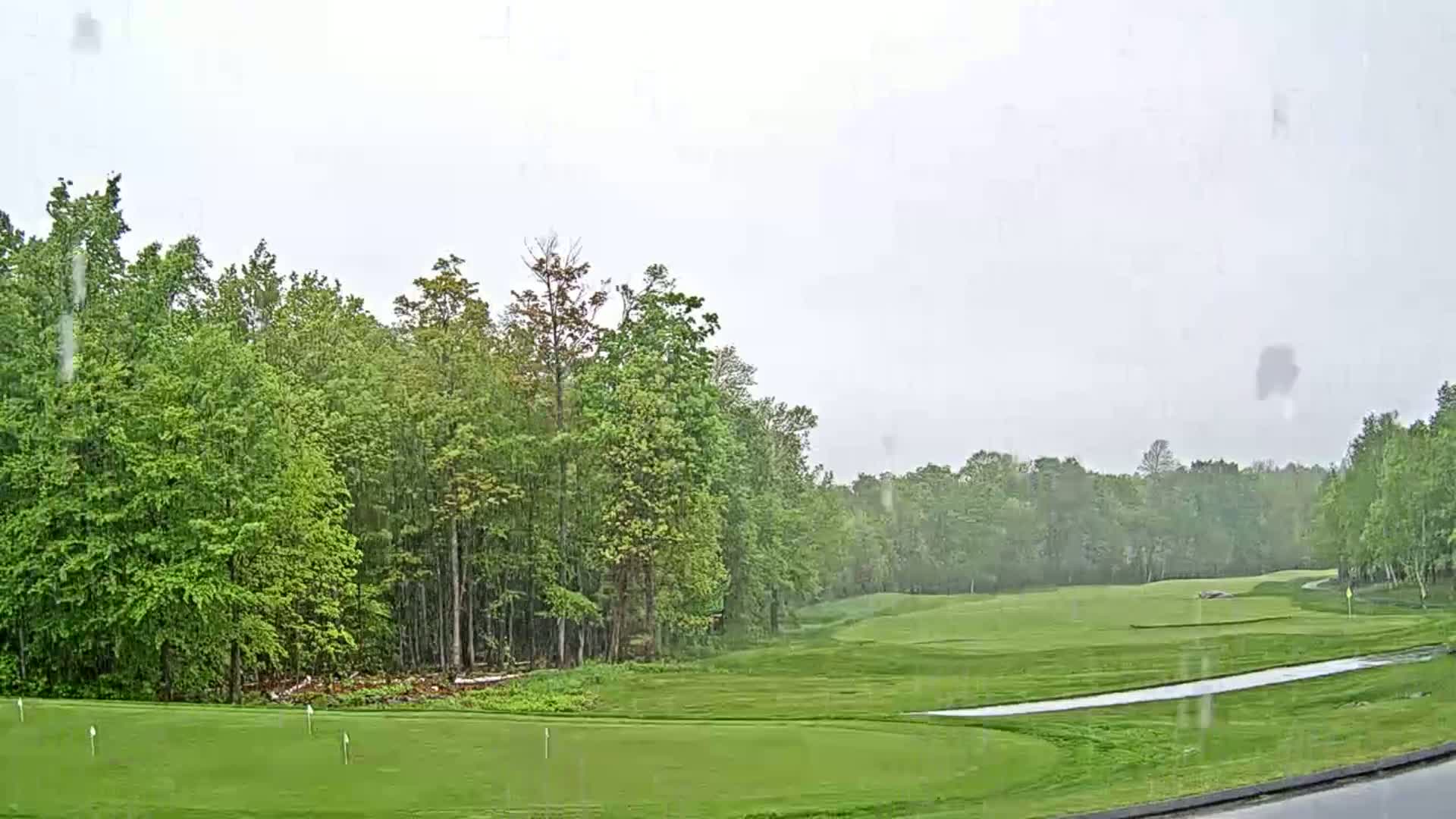 A golf course surrounded by lush green trees is seen during a light rain.