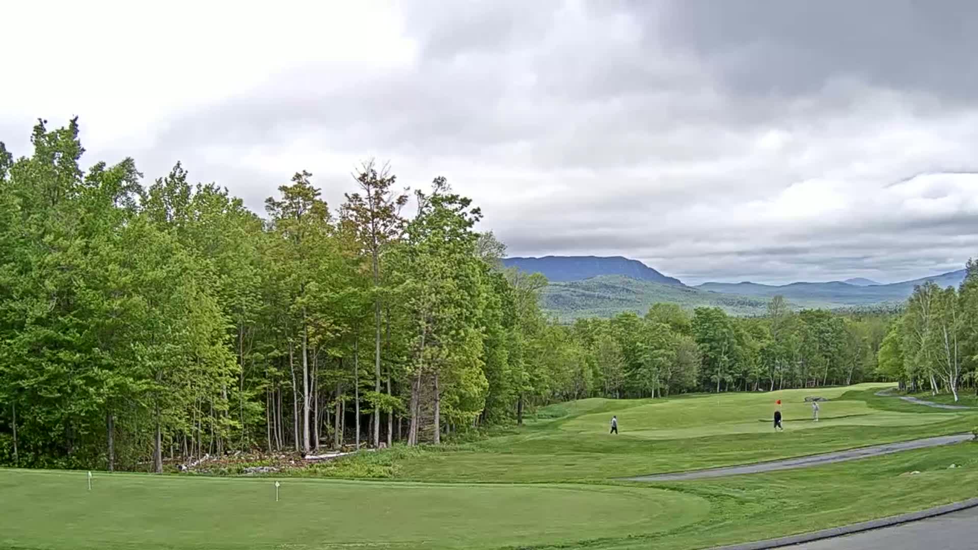 A golf course with green fairways and lush trees, under a mostly cloudy sky, overlooks a distant mountain range.