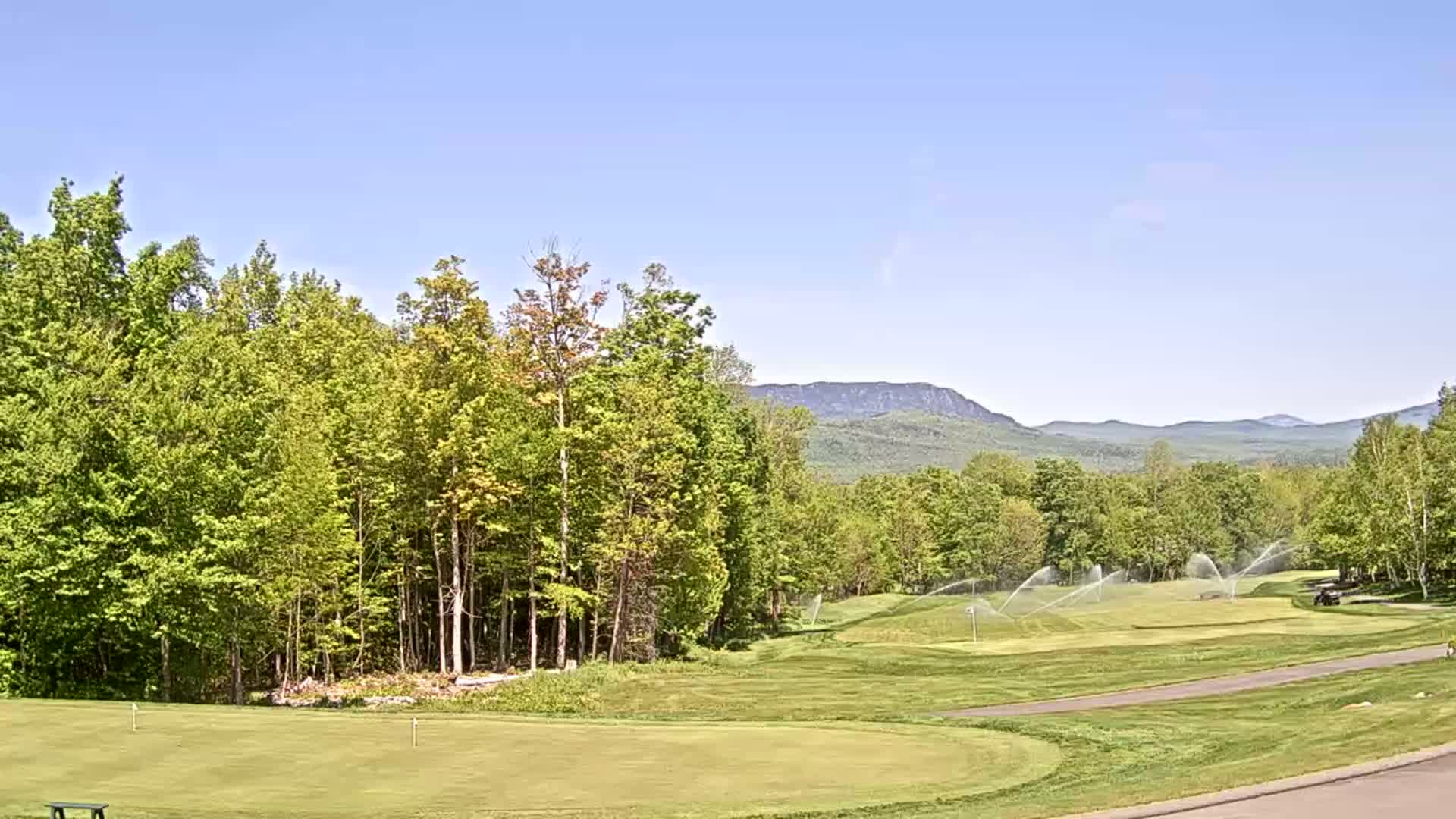 A golf course with sprinklers spraying water, surrounded by lush green trees and mountains under a clear blue sky.