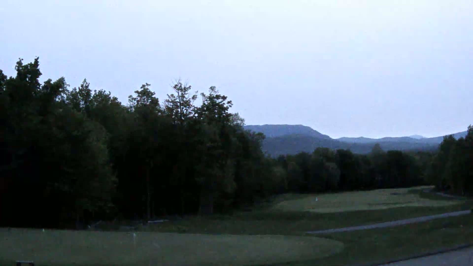 A golf course is situated in front of a line of dark trees, with hazy mountains visible in the background under a pale, overcast sky.