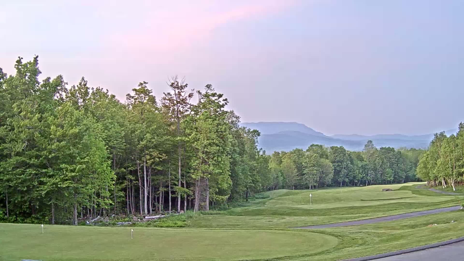 A golf course with green fairways and a cart path, bordered by lush green trees, under a pale, hazy sky with distant, hazy mountains in the background.