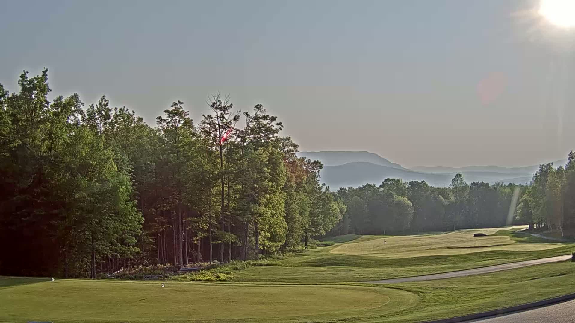 A sunny, hazy day reveals a golf course nestled among lush green trees, with distant mountains visible on the horizon.