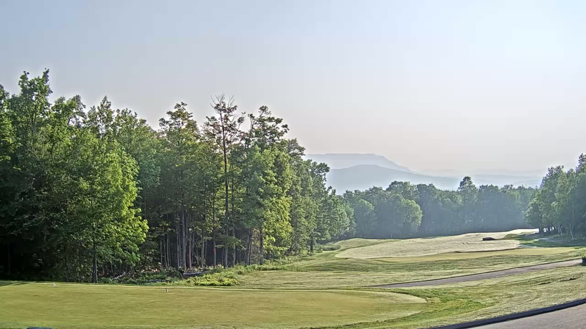 A golf course fairway stretches towards hazy mountains under a clear sky.
