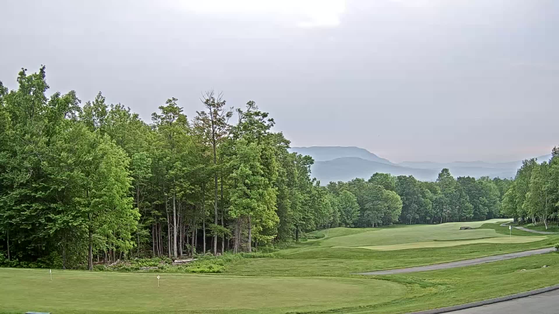 A golf course green and fairway are situated amidst lush green trees, with hazy mountains visible in the distance under an overcast sky.