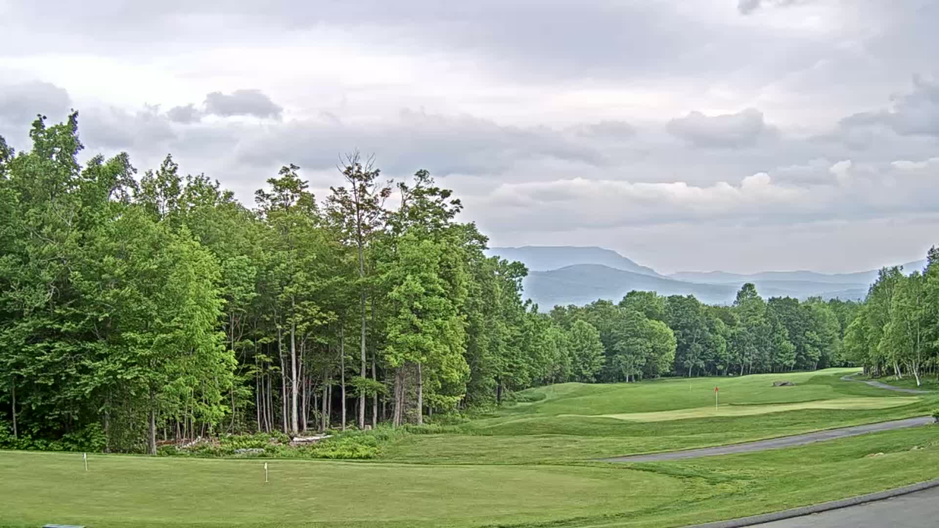 A golf course with lush green fairways and trees under a cloudy sky, mountains visible in the distance.