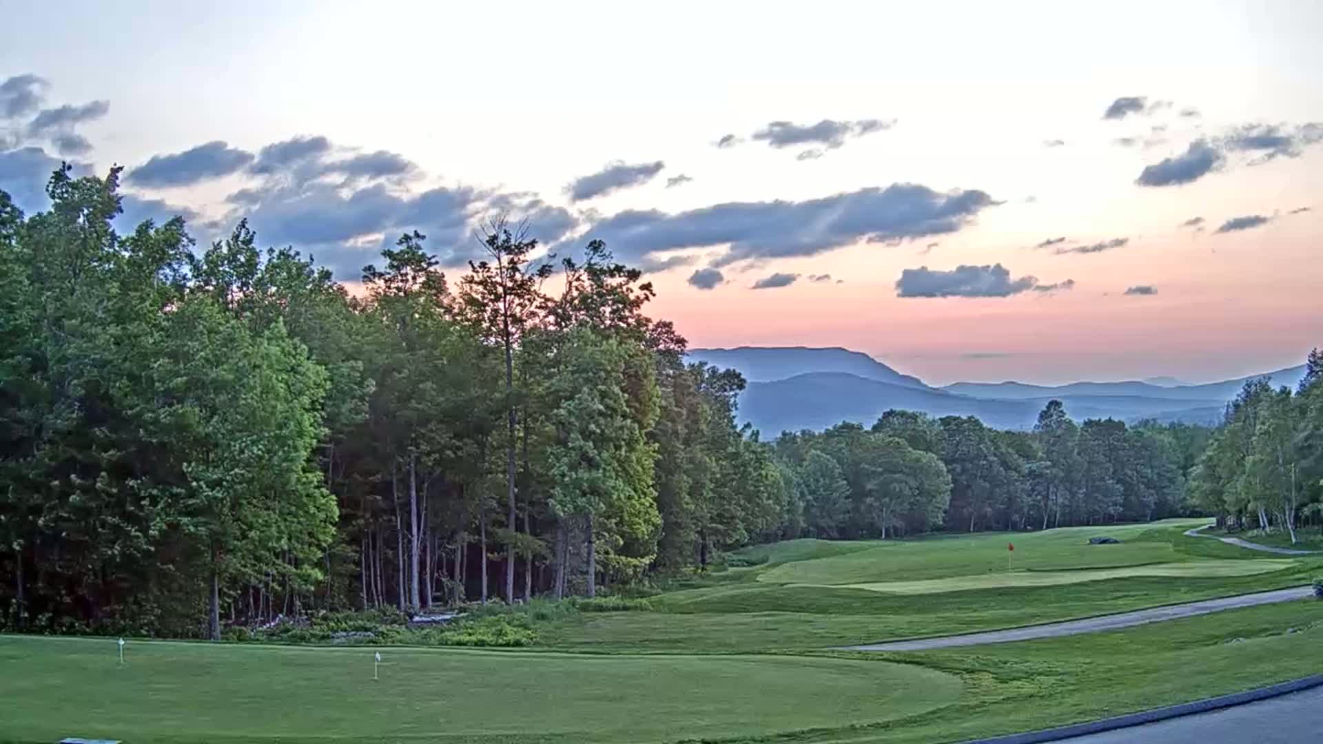 A golf course with green fairways and lush trees overlooks a range of mountains under a pastel sunset sky.
