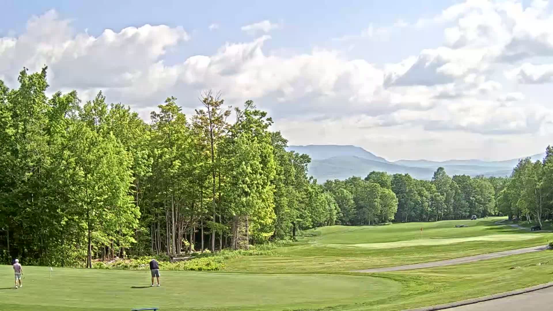 A partly cloudy day on a golf course nestled among green trees, with distant blue mountains visible in the background.