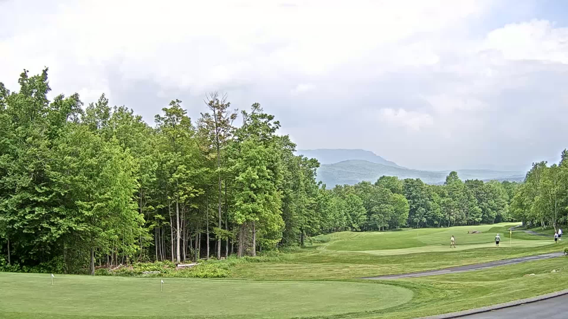 A golf course with green fairways and putting greens, surrounded by lush green trees and a hazy mountain range in the background under a partly cloudy sky.