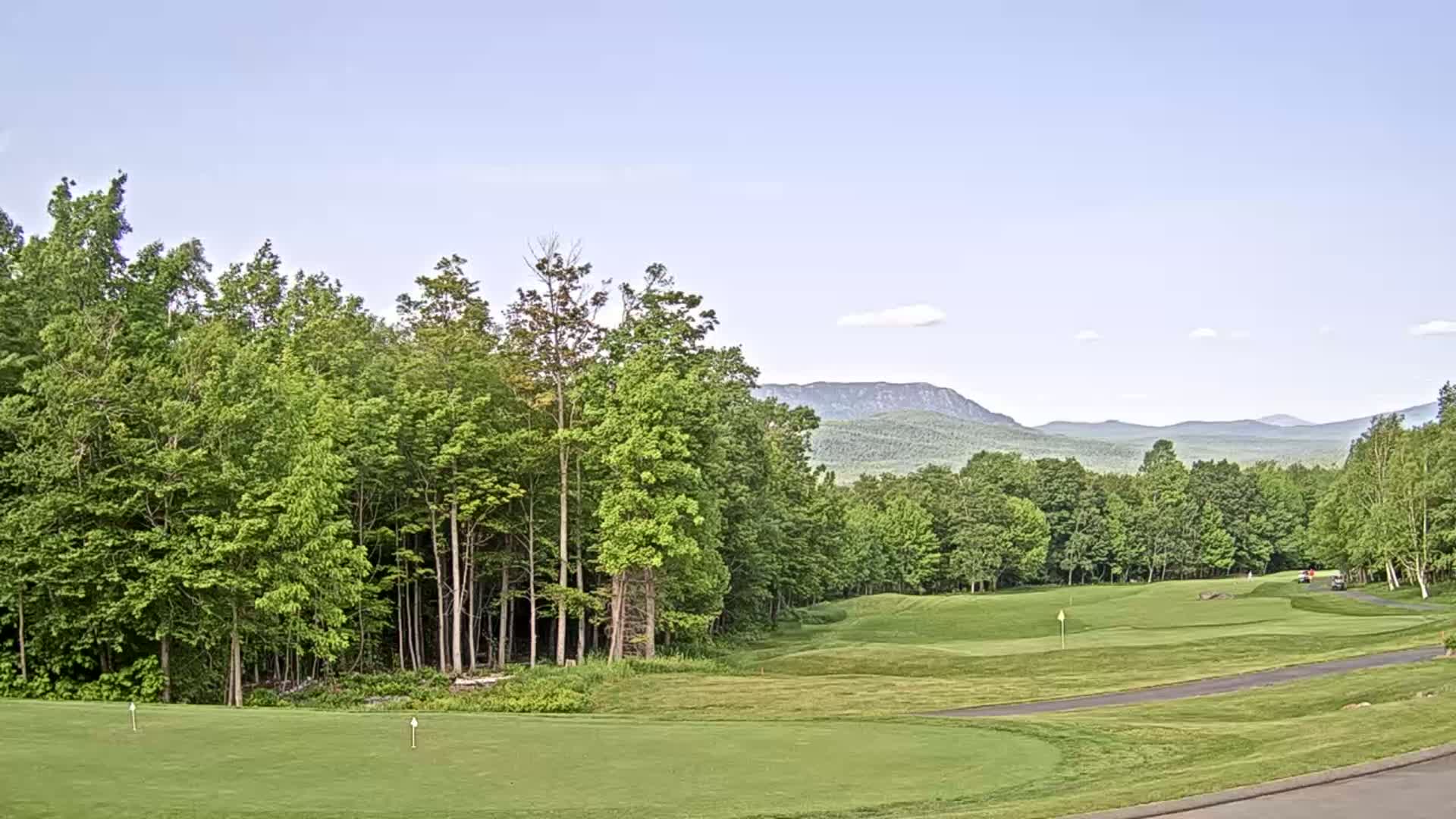 A golf course fairway and green, bordered by lush green trees and a mountain range under a clear, light blue sky.