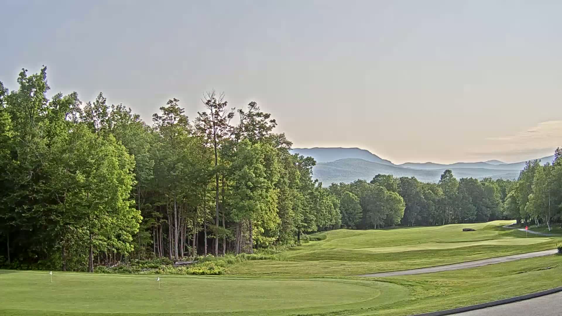 A golf course green, bordered by lush green trees and a distant mountain range under a clear, light sky.