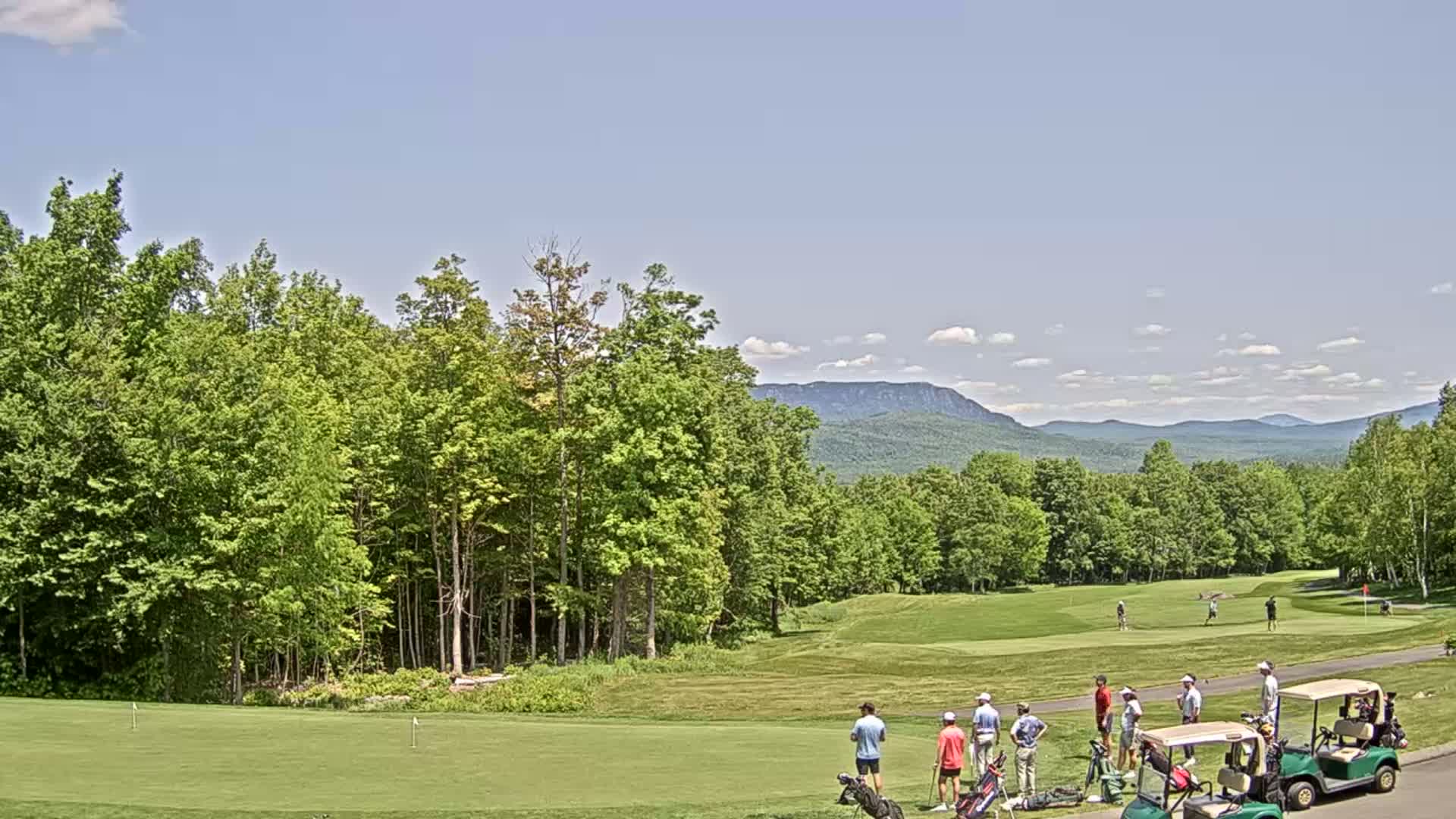 A golf course with several golfers and golf carts on a putting green, surrounded by lush green trees and mountains under a mostly clear sky.