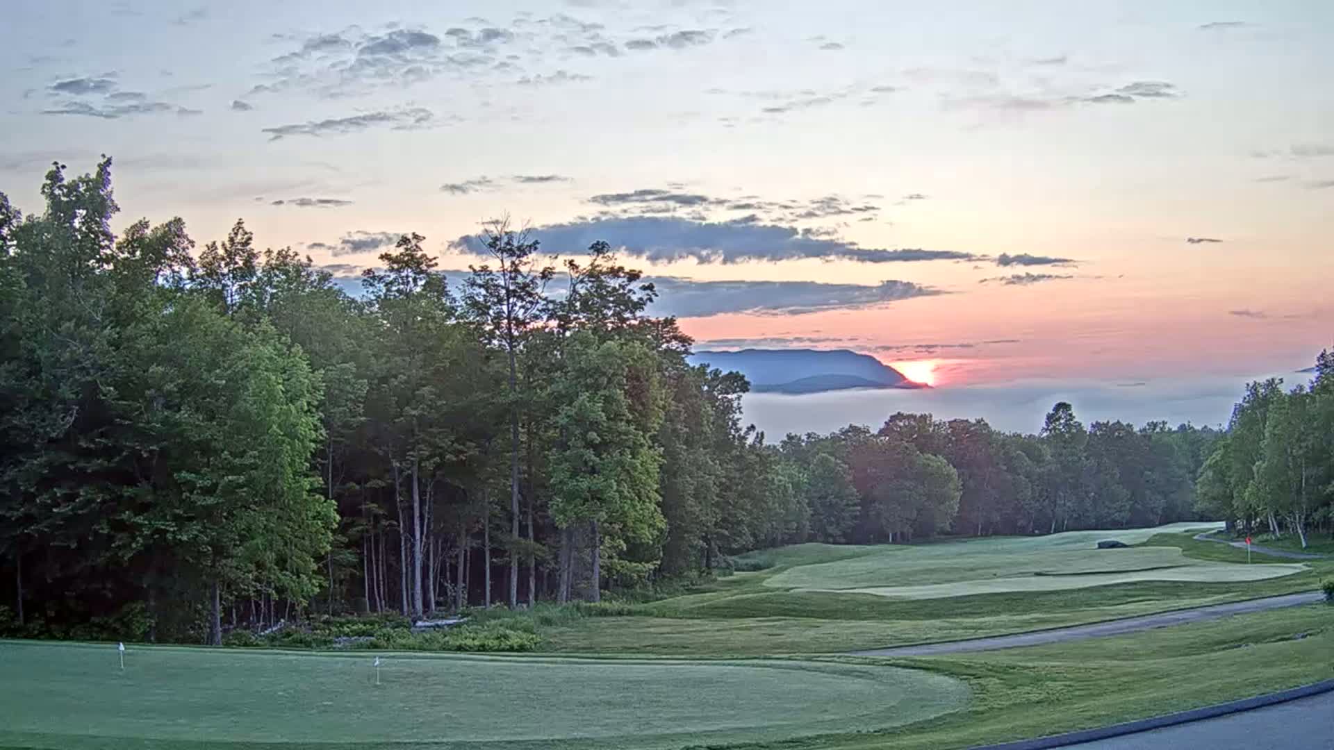A golf course nestled in a forest overlooks a valley filled with fog at sunrise under a pastel sky.