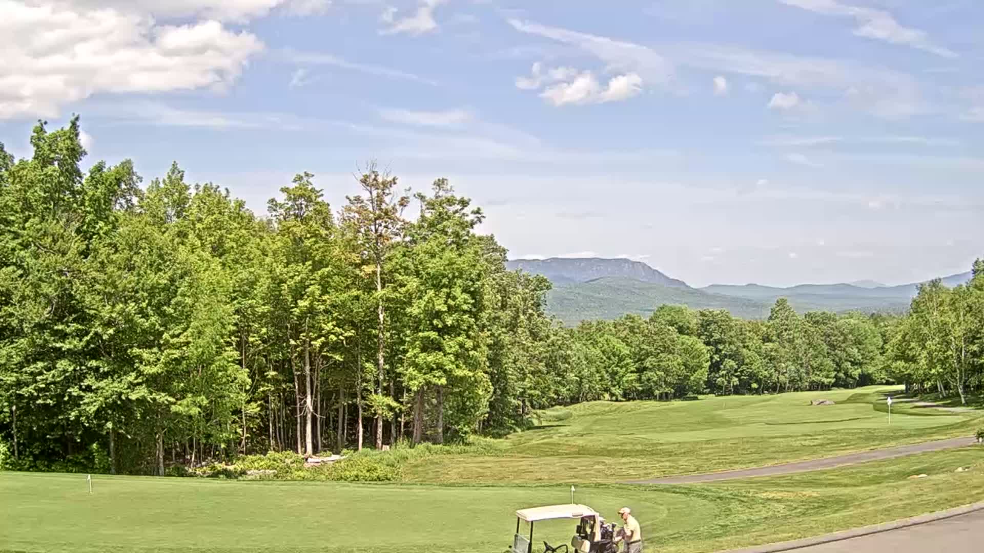 A golf course with a golf cart and two people on it, surrounded by lush green trees and rolling hills under a partly cloudy sky.
