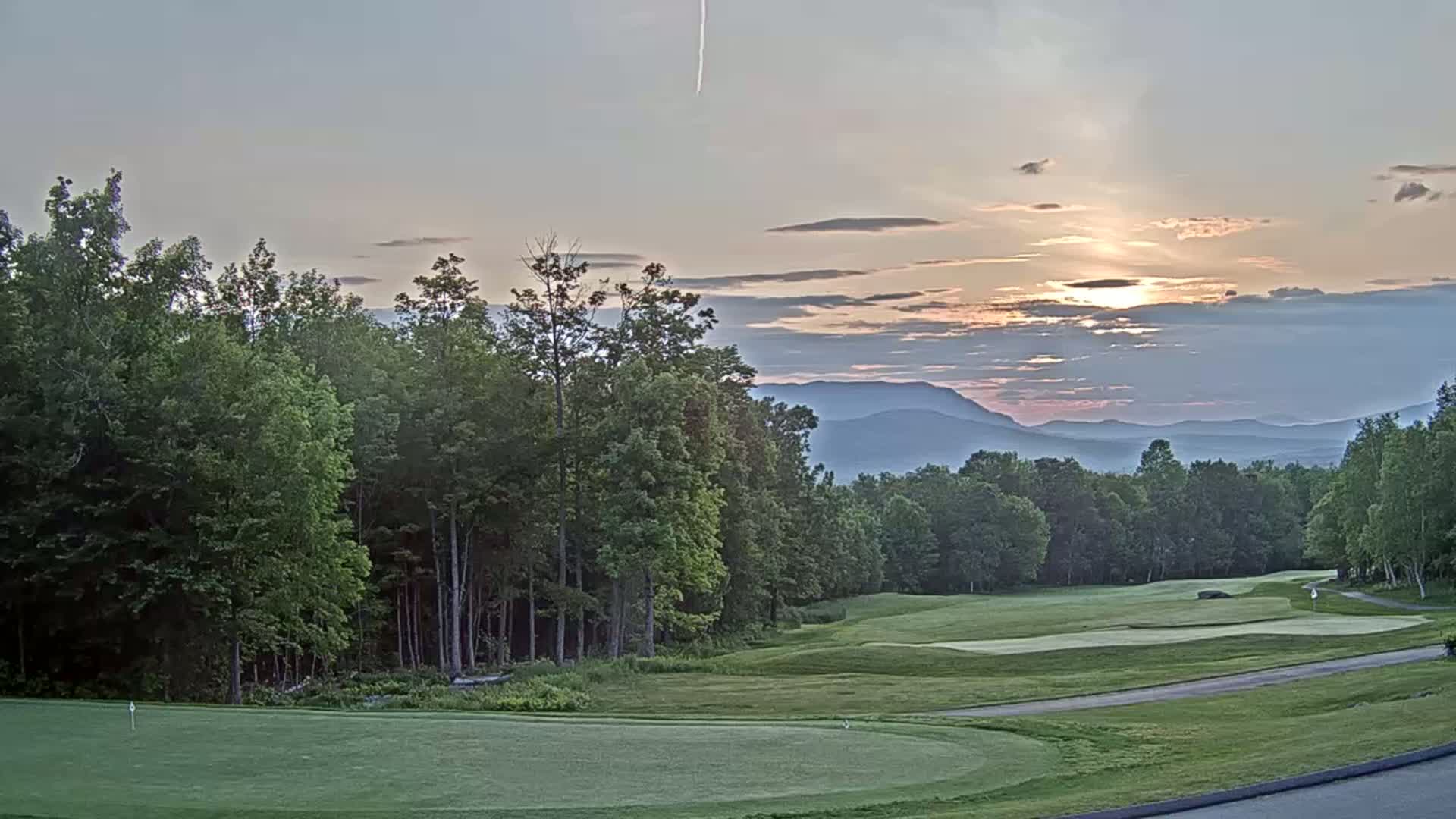 A golf course with a green fairway and putting green is nestled among lush green trees, overlooking a mountain range under a partly cloudy sunset sky.