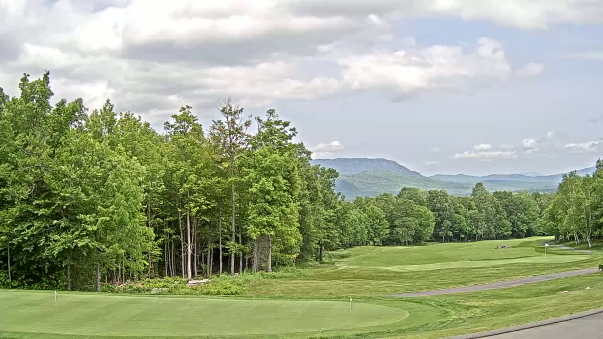 A partly cloudy day reveals a golf course nestled among lush green trees and distant mountains.