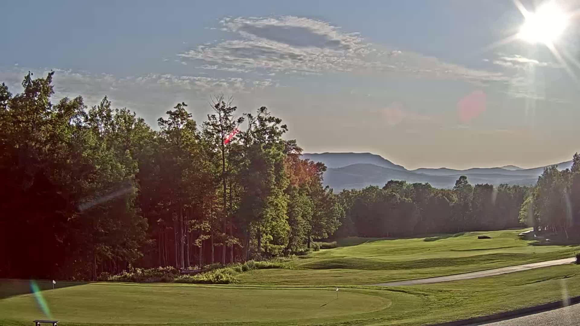 A sunny day reveals a golf course nestled among green trees, with distant blue mountains under a partly cloudy sky.