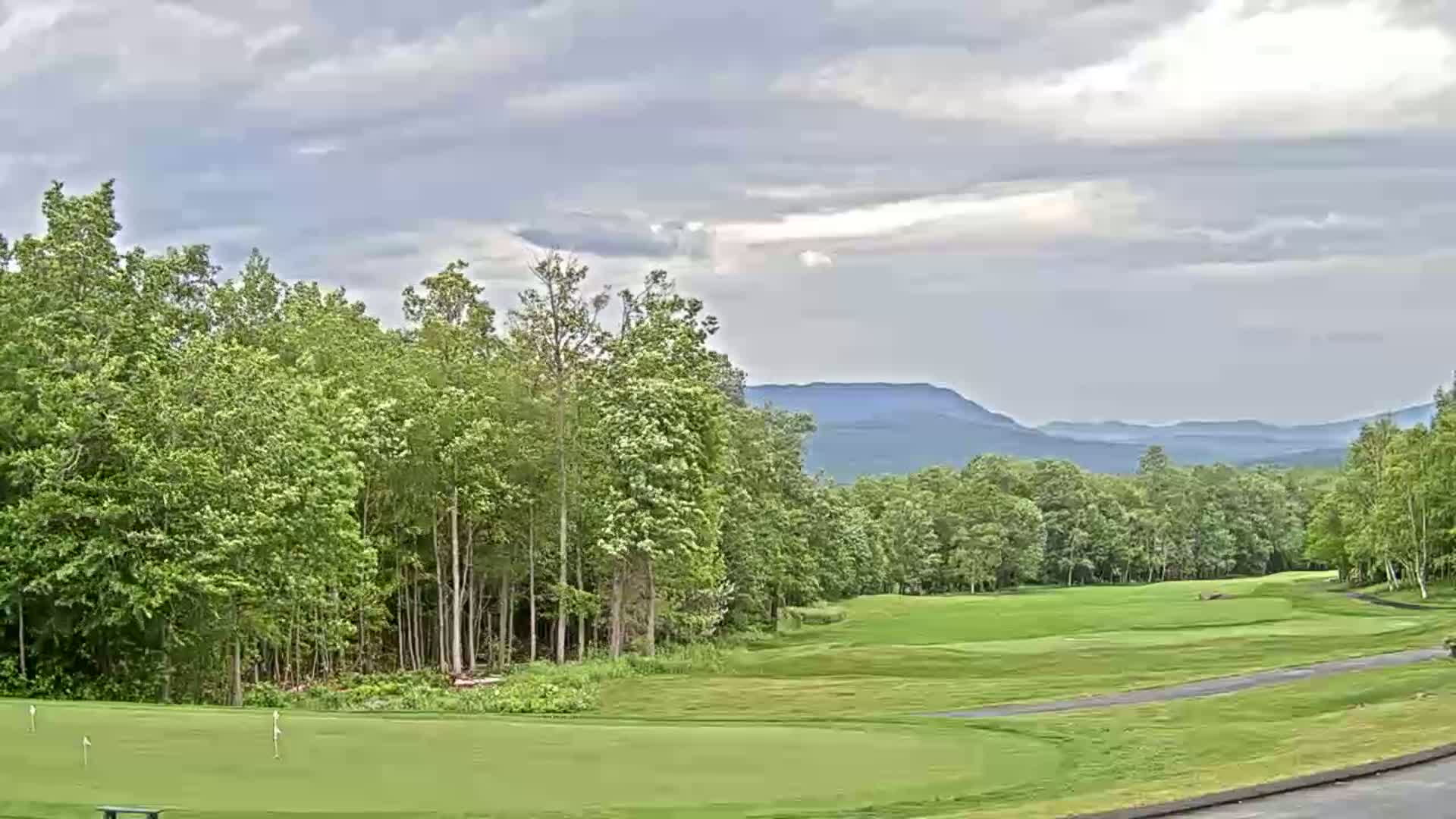 A golf course with green fairways and lush trees, under a partly cloudy sky, overlooks distant mountains.