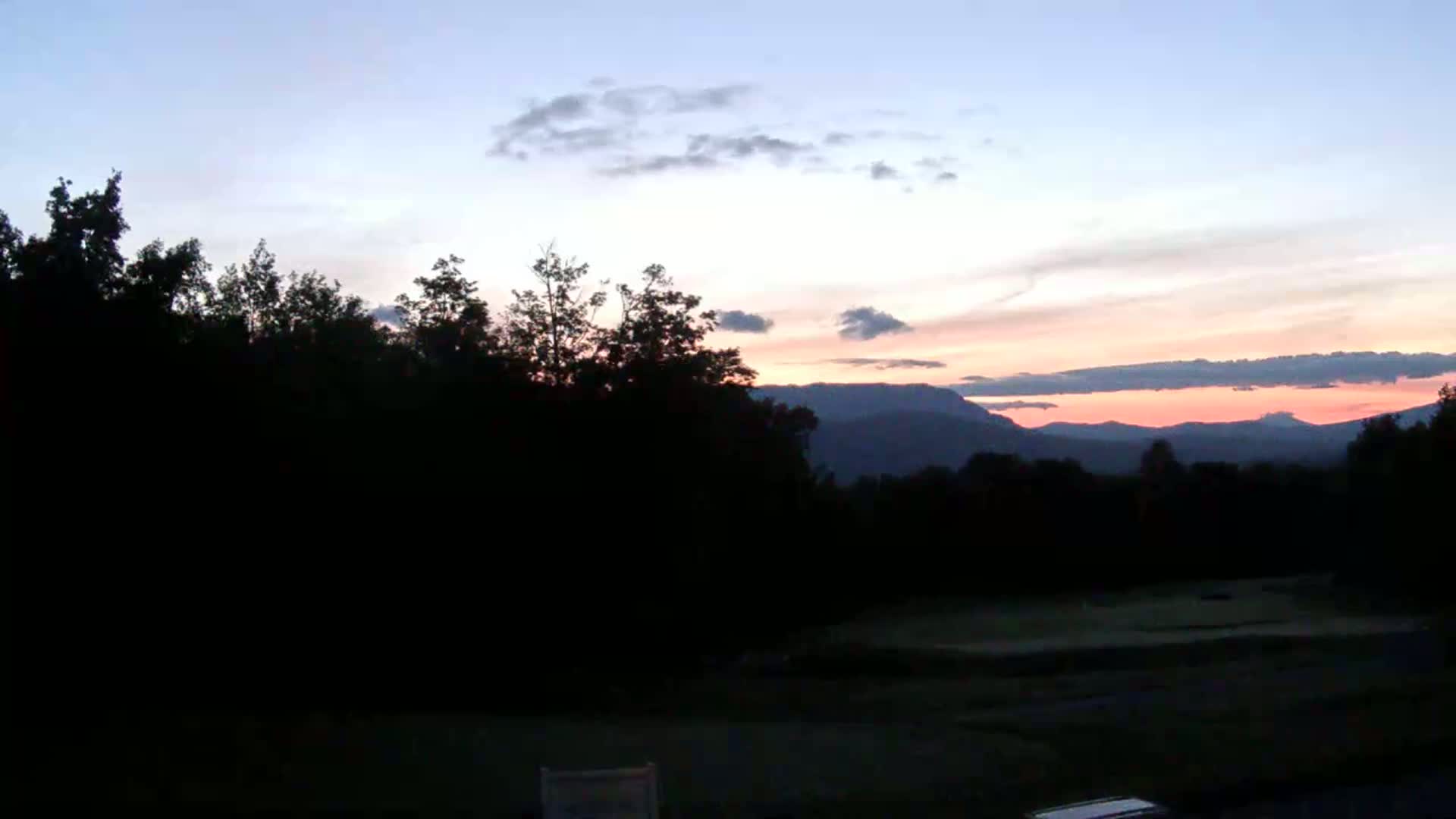 Silhouetted trees frame a sunset over distant mountains under a partly cloudy sky.