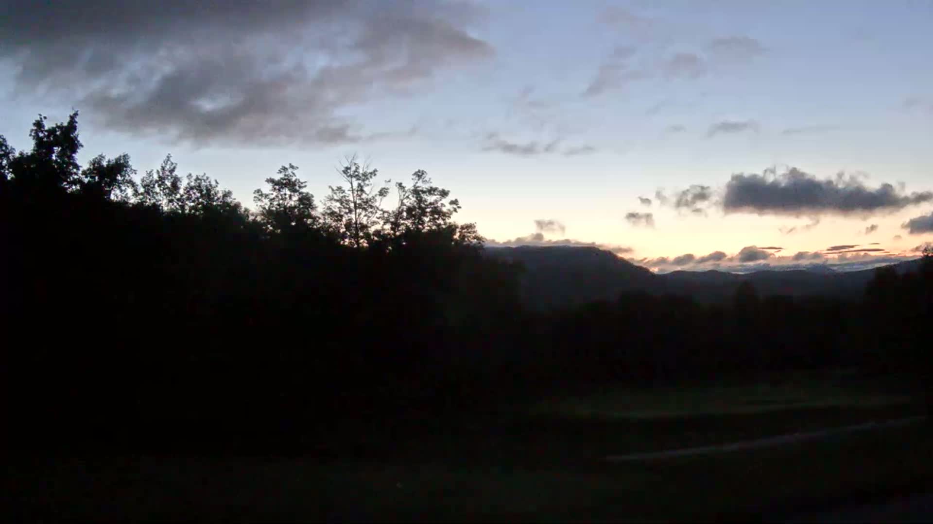 Silhouetted trees stand in the foreground against a partly cloudy twilight sky, with distant mountains visible on the horizon.