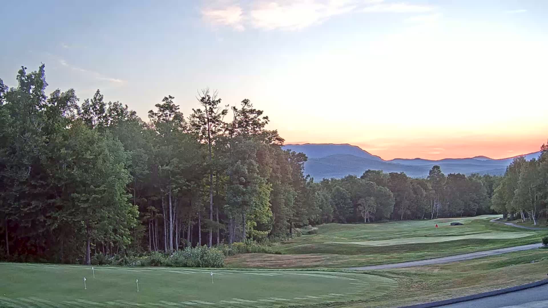 A putting green on a golf course bordered by lush green trees, with a mountain range visible in the background under a cloudy sky.