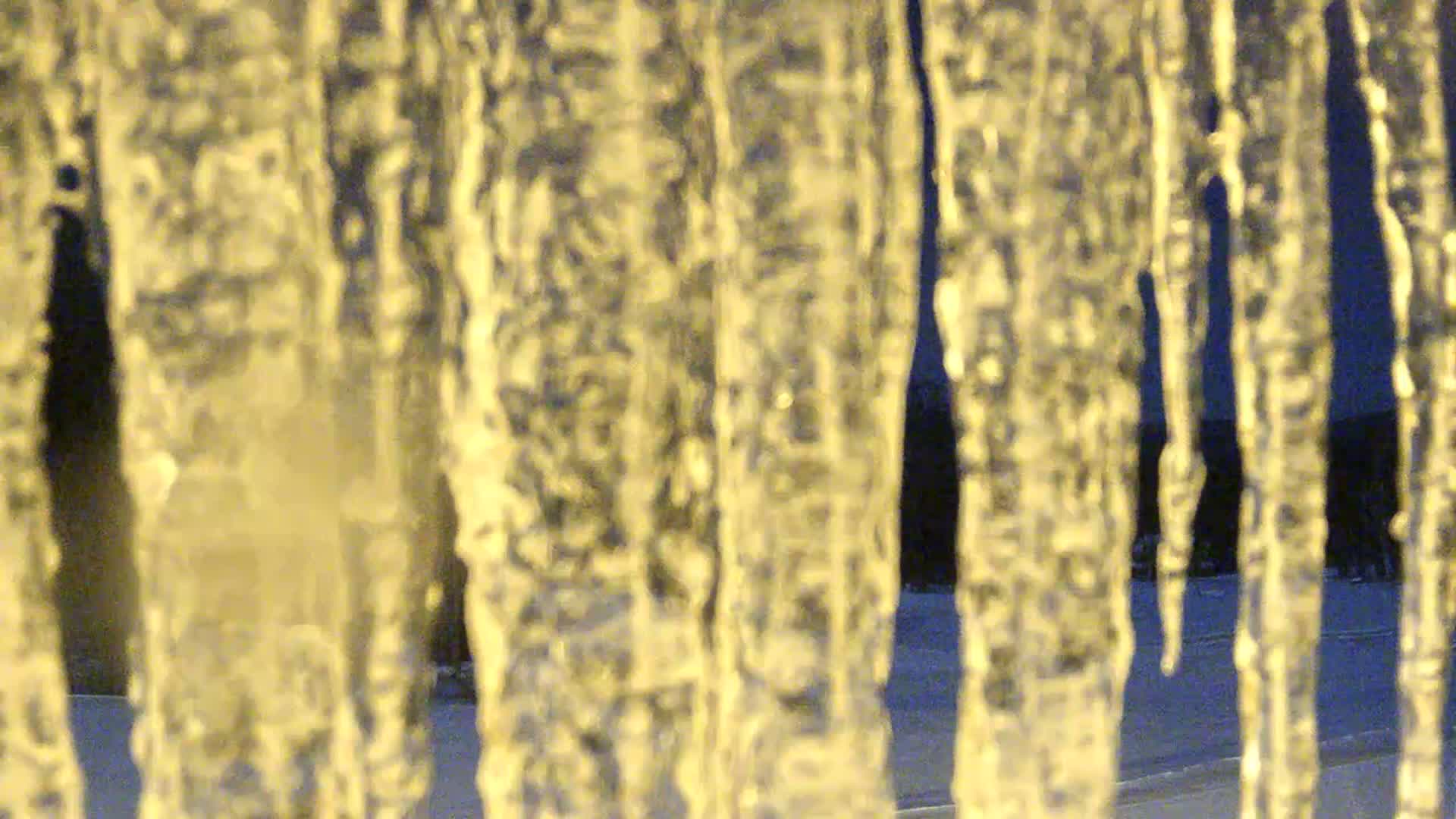 Numerous golden-hued icicles hang closely in the foreground against a blurred, dark blue background, indicating cold and freezing outdoor conditions.