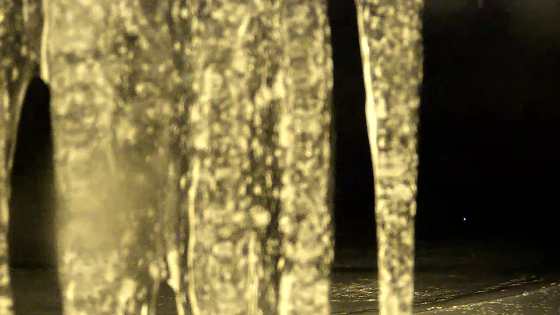 Several blurry, golden-hued icicles hang prominently in the foreground against a dark winter night, with a hint of snow on the ground, indicating freezing outdoor temperatures.