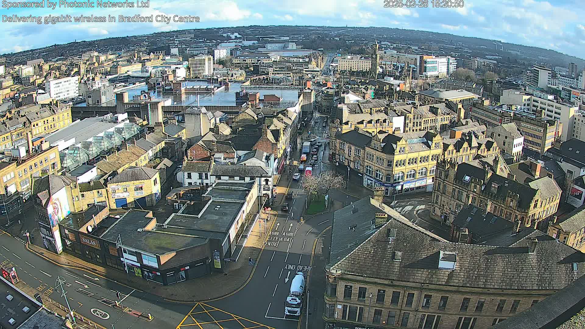 A high-angle, long shot of a city center on a partly sunny day shows many buildings, streets, and vehicles.