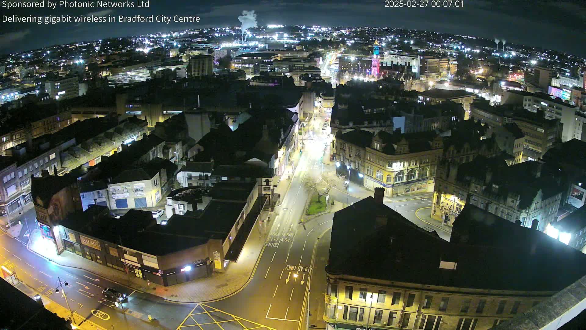A nighttime aerial view of a city center, showing illuminated streets and buildings under a partly cloudy sky.