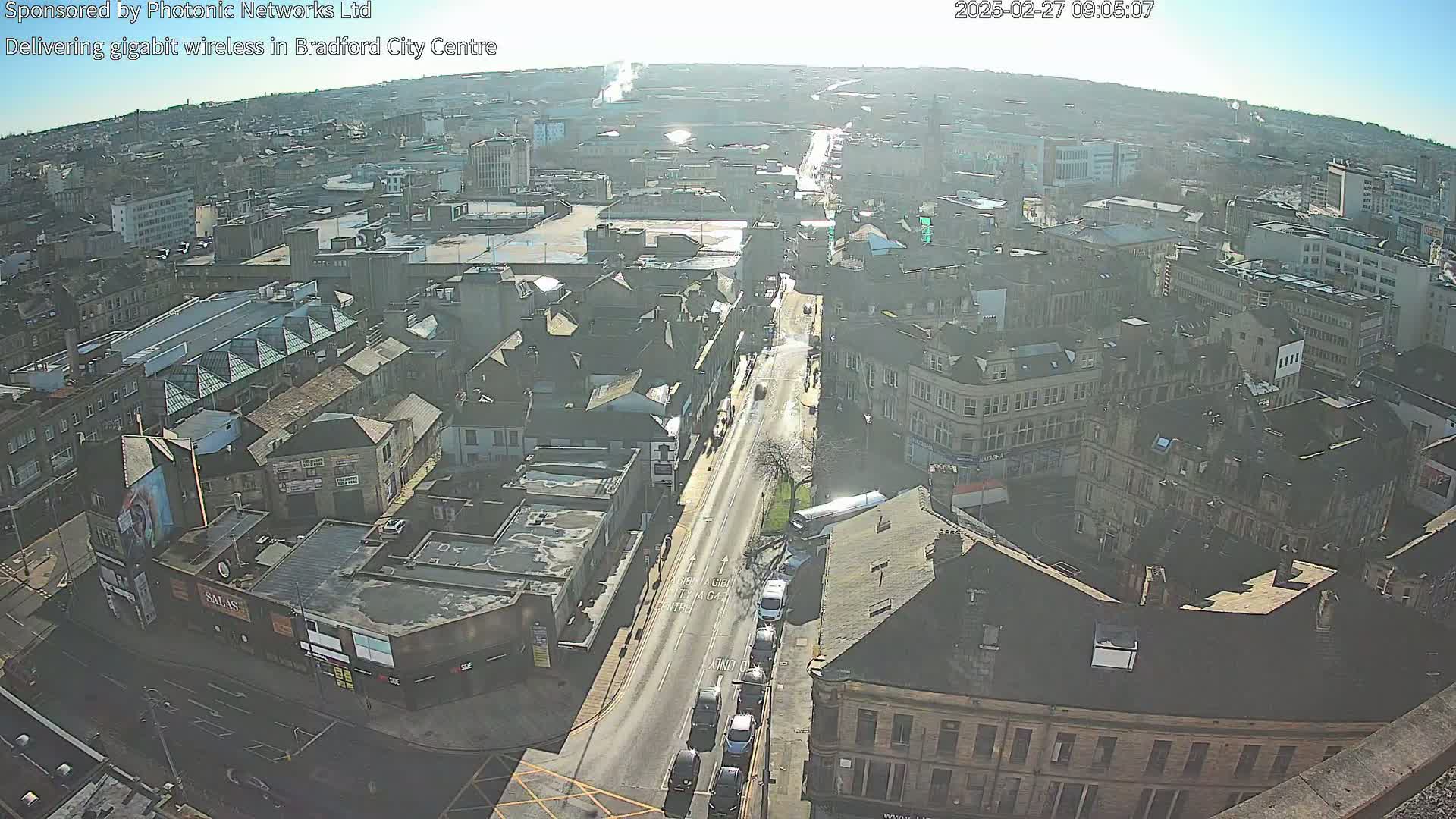 A high-angle, long shot of a city street on a sunny day, showing numerous buildings and some vehicles on the road.