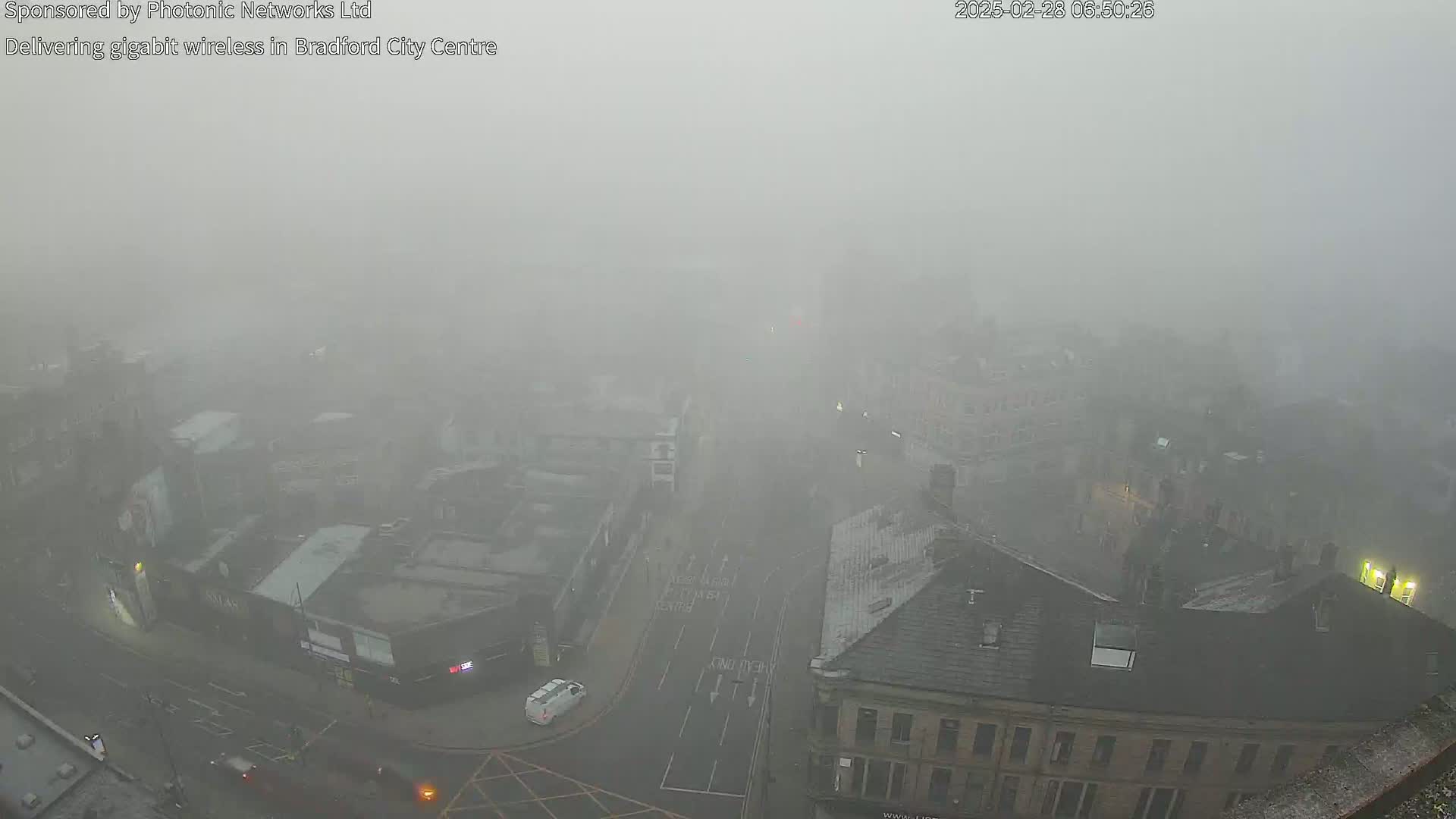 A thick fog obscures a city street scene, with a few vehicles visible on a road intersection amongst numerous buildings.