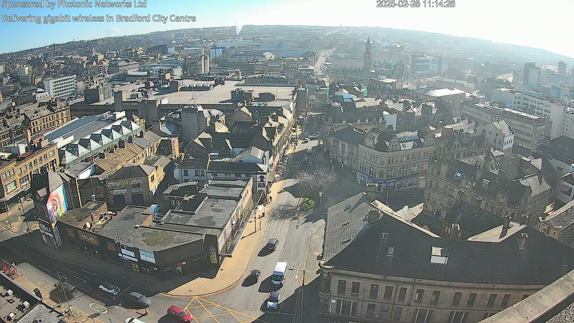 A high-angle, long shot of a city center on a sunny day shows numerous buildings, streets, and a few vehicles.