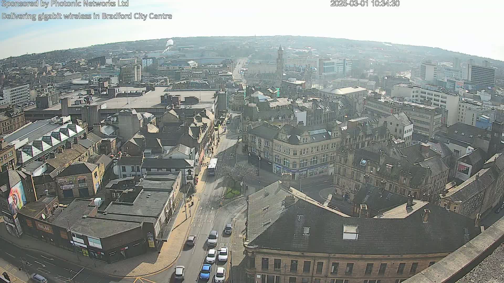 A high-angle, long shot on a sunny day shows a city center with many buildings and a few cars on the roads.